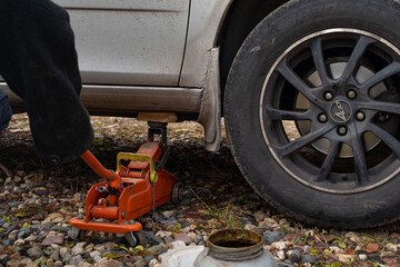 Fototapeta premium Jack. A white car is lifted by a car jack and a pair of wheels on a dirt road. The car is lifted by a jack. Car wheel repair. Part of the photo is blurred.