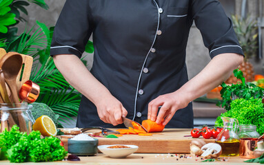 Chef cuts carrots into wheels with santoku knife. Cozy kitchen with wooden table, kitchenware, vegetables, herbs and ingredients for cooking. Healthy vegan food, culinary, recipes, food blogging