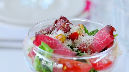 Glass bowl displaying mixed salad featuring sliced seasoned meat, ripe tomatoes, grated cheese, and fresh herbs, resting on crisp white tablecloth