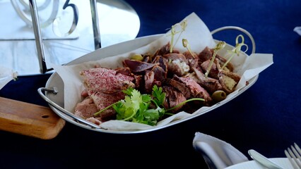 Metal tray displaying sliced charcuterie, rolled meats adorned with fresh herbs, olives, accompanied by numbered identification card on rustic surface