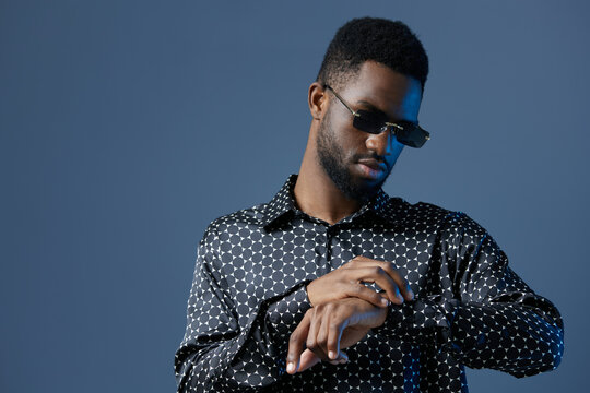 Stylish man in sunglasses and polka dot shirt checking time on his watch against a cityscape backdrop