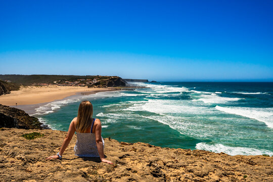 Beautiful mature woman sitting on cliff overlooking ocean and sandy beach Monte Clerigo, Algarve, Portugal on sunny day. Back view