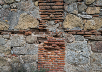 Rustic wall made of irregularly shaped stones and red bricks. The stones are mostly gray and beige,...