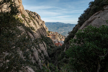 Obraz premium Views of the Monastery of Montserrat, Barcelona, Spain, from the Santa Maria gorge, surrounded by rugged rocky cliffs and dense vegetation, with distant mountains under a clear blue sky