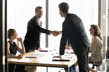 Two millennial businessmen shake hands standing at conference table, establish strategic alliance...