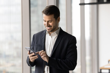 Smiling millennial businessman dressed in suit using modern smartphone standing in office, solving business-related matter by e-mail, making call, download new business application, chatting, texting