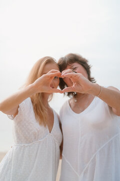 Young red haired woman and her senior mother on vacation on the beach by the ocean put their hands together in a heart and smile. Symbol of love and family. Mother's Day.