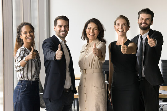 Diverse group of five businesspeople standing together, giving thumbs-up gesture to camera, sign of approval, successful teamwork, professional achievement, sense of unity, and effective collaboration