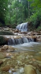 cascading waterfall, surrounded by lush greenery and moss-covered rocks, creates an enchanting natural scene. The water is crystal clear with soft ripples as it flows down the rocky surface