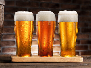 Three glasses of chilled beer on old wooden table and brick wall at the background.