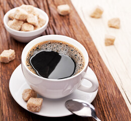 Cup of coffee on white plate and brown sugar cubes on wooden table close up. Top view.