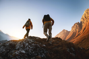 Silhouettes of two active sporty young hikers walks with backpacks high in mountains against sunset...