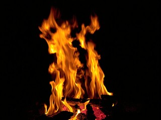 Bright orange and yellow flames of a campfire dancing against a dark night sky, with glowing embers
