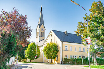 Sunny street view of church with tall steeple and surrounding trees in Weichering, Bavaria, Germany
