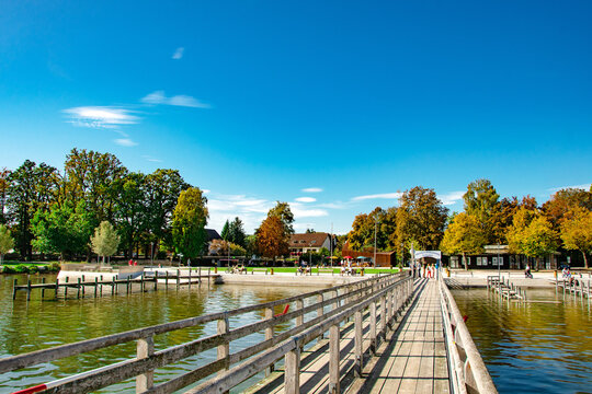 Peaceful lake landscape with wooden pier, sailboats and trees under clear blue sky
