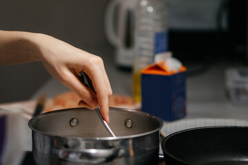 A close-up shot of a hand stirring food in a stainless steel pot on a stovetop, with a blurred background of kitchen ingredients and utensils, capturing a cozy home cooking moment