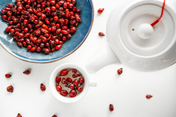 Composition of pouring rose hips herbal tea from a white teapot into a cup and dried red berries on an around blue ceramic plate on a white wooden table. Rustic style. Immune system support. Top view.
