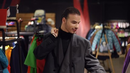 A confident fashion consultant in a black turtleneck and blazer carries colorful garments over his shoulder while browsing stylish clothing in a boutique. His poised expression and elegant attire