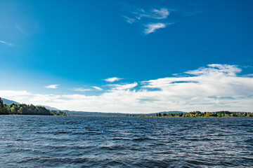 Obraz premium Scenic view of Lake Ammersee with a calm water surface, surrounded by lush greenery and distant mountains under a bright blue sky.