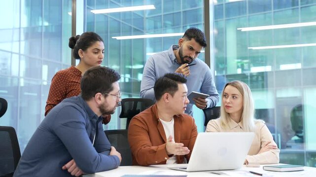 Manager explains startup project details to group of workers using laptop sitting at workplace in modern business office. Businessman discussing with colleagues, collaboration, communicating teamwork