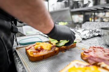 A close-up of a chef's hand in a black glove preparing a sandwich. Fresh ingredients, including...