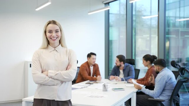 Portrait of a smiling business woman on background diverse team employees at workplace in a office. Confident optimistic female worker posing with crossed arms looking at camera near group colleagues