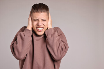 Annoyed young man covering his ears due to loud sound on light grey background