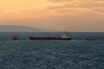A large red cargo ship is escorted by a tugboat on a calm sea during a beautiful sunset, with distant mountains visible on the horizon.