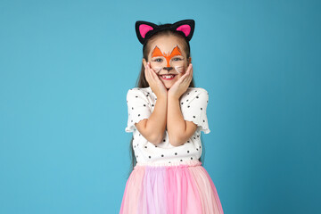 Cute girl with painted face and ears as cat on light blue background