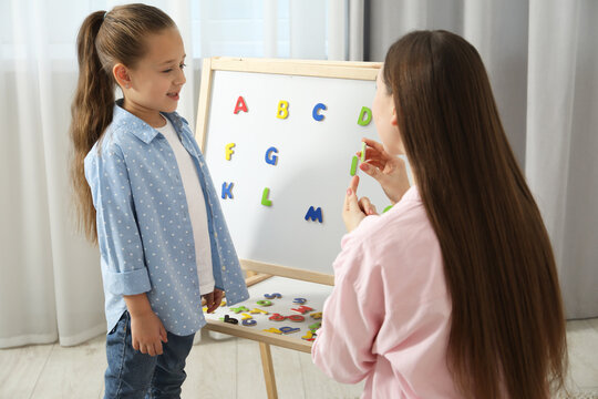 Speech therapist teaching little girl alphabet with magnetic letters indoors