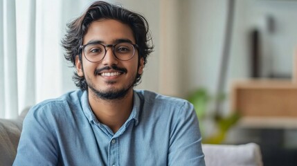 Fototapeta premium A cheerful young Indian man enjoys a moment of relaxation on a soft sofa, exuding warmth and positivity. The stylish living area is bathed in natural light, creating a vibrant atmosphere