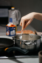 A close-up shot of a hand stirring food in a stainless steel pot on a stovetop, with a blurred background of kitchen ingredients and utensils, capturing a cozy home cooking moment