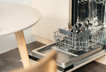 Open dishwasher in beige design made of furniture plywood with basket and forks in the kitchen near the table. technologies that make life easier.