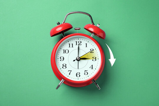 Daylight saving time. Red alarm clock on green background, top view. Highlighted area and arrow showing time change by one hour forward
