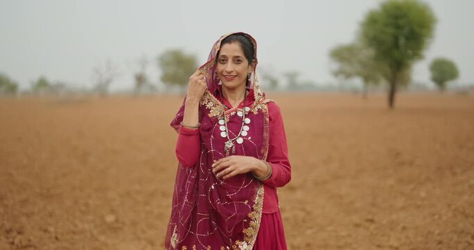 Indian young adult village farmer woman wear red saree standing outdoor farmland looking at camera. Asian happy smiling mother do pose staring cam outside ground