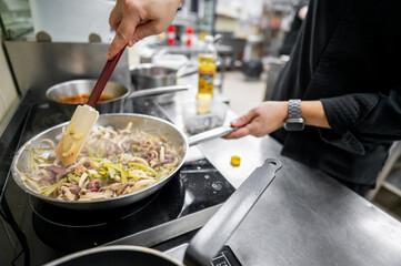 A chef stirs vegetables and meat in a skillet on a stove in a professional kitchen, showcasing culinary skill and fresh ingredients. The scene captures the essence of food preparation.