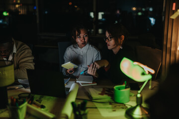 A diverse group of colleagues collaborates intensely around a desk in a dimly lit office. They are engaged in brainstorming sessions and analyzing project details, aiming to meet an upcoming deadline.