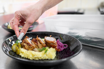 A close-up of a gourmet plate featuring sliced chicken, creamy mashed potatoes, pickles, and red onions. A hand is delicately adding a garnish, enhancing the dish's presentation.