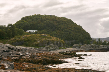 White house perched on rocky hillside with dense forest and cloudy sky.