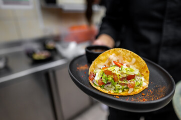 A close-up of a freshly prepared taco served on a black plate. The taco is filled with lettuce, tomatoes, and spices, accompanied by a small bowl of sauce. Ideal for food-related content.