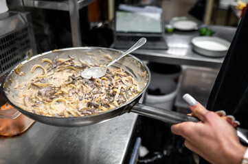 A chef holds a pan of creamy mushroom dish, steaming and ready to serve. The kitchen setting features stainless steel equipment and a busy atmosphere, highlighting culinary preparation.