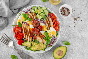 Healthy cobb salad with roasted chicken breast, chopped avocado, tomatoes, bacon, cucumber, hard boiled egg and green leaves on a plate. Top view.