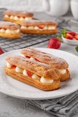 Eclairs with cream and fresh strawberries sprinkled with powdered sugar on top on a gray concrete background.