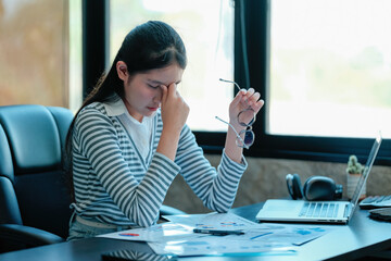 Young Asian businesswoman suffering from headache while using laptop computer working in modern...