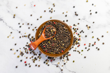 A wooden bowl filled with mixed peppercorns, including black, red, green, and white varieties, placed on a marble surface. A wooden spoon rests on top, adding a rustic touch to the culinary scene.
