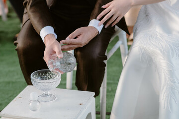 Groom wear a brown suit and the bride wear a white embroidered dress. Pouring water for auspiciousness on the wedding day, close up, two people
