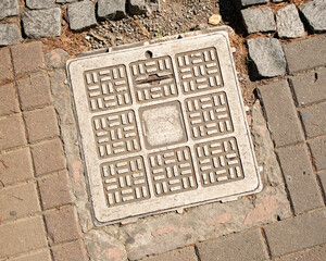 Manhole cover with intricate design and local inscriptions located on a city sidewalk during daylight hours. 