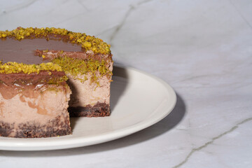 Close up Dubai chocolate cheesecake with pistachio paste and kataifi covered in chocolate ganache on white plate. White marble background with copy space. Indulgence, gourmet desserts, art of culinary