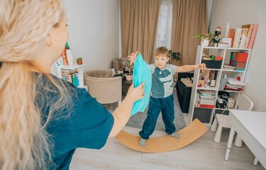 boy developing motor skills and coordination by playing on a wooden wobble board during a therapy session, interacting with a therapist