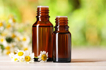 Bottles of essential oil and chamomile flowers on white wooden table against blurred background, closeup